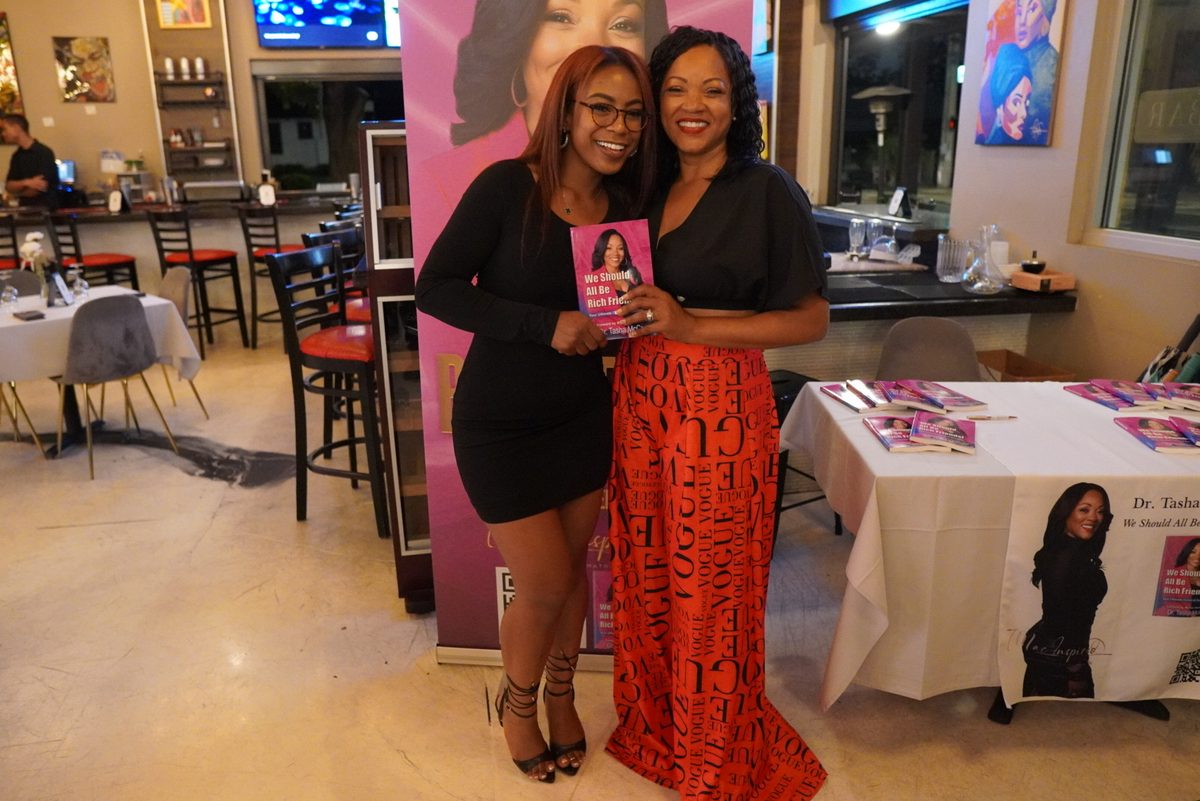 A reader in red scrubs holding the book beside the tour banner